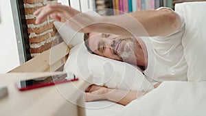 Middle age man lying on bed turning off smartphone alarm at bedroom