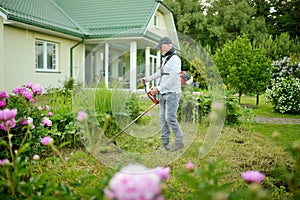 Middle age man mowing tall grass with electric or petrol lawn trimmer in a backyard. Gardening care tools and equipment