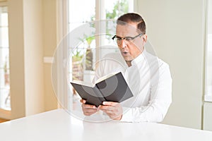 Middle age man reading a book at home wearing glasses scared in shock with a surprise face, afraid and excited with fear