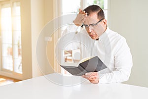 Middle age man reading a book at home wearing glasses stressed with hand on head, shocked with shame and surprise face, angry and