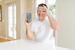 Middle age man showing smartphone screen with happy face smiling doing ok sign with hand on eye looking through fingers