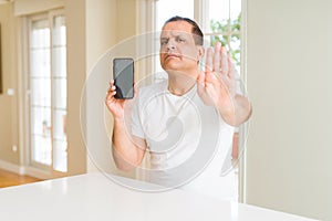 Middle age man showing smartphone screen with open hand doing stop sign with serious and confident expression, defense gesture