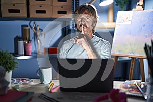 Middle age man sitting at art studio with laptop at night asking to be quiet with finger on lips