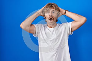 Middle age man standing over blue background crazy and scared with hands on head, afraid and surprised of shock with open mouth