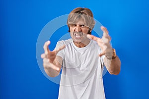 Middle age man standing over blue background shouting frustrated with rage, hands trying to strangle, yelling mad