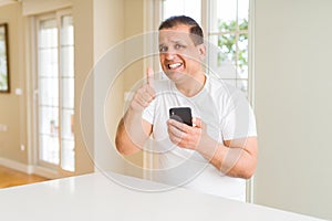 Middle age man using smartphone at home happy with big smile doing ok sign, thumb up with fingers, excellent sign