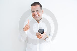 Middle age man using smartphone over white wall happy with big smile doing ok sign, thumb up with fingers, excellent sign