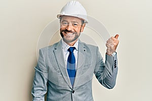 Middle age man wearing architect hardhat pointing thumb up to the side smiling happy with open mouth