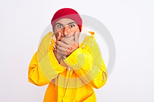 Middle age man wearing rain coat and woolen hat standing over isolated white background shocked covering mouth with hands for
