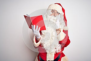 Middle age man wearing Santa Claus costume reading book over isolated white background afraid and shocked, surprise and amazed