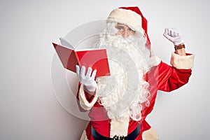 Middle age man wearing Santa Claus costume reading book over isolated white background Strong person showing arm muscle, confident