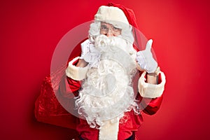 Middle age man wearing Santa costume holding sack with gifts over isolated red background happy with big smile doing ok sign,