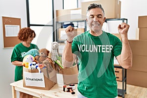 Middle age man wearing volunteer t shirt at donations stand excited for success with arms raised and eyes closed celebrating