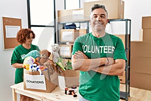 Middle age man wearing volunteer t shirt at donations stand happy face smiling with crossed arms looking at the camera