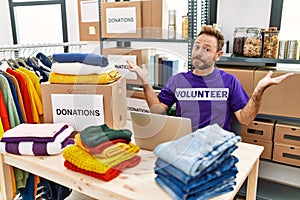 Middle age man wearing volunteer t shirt working with laptop clueless and confused expression with arms and hands raised