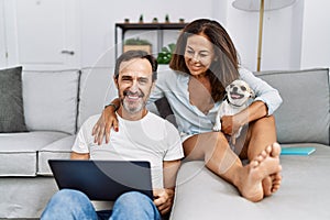 Middle age man and woman couple using laptop sitting on sofa with chihuahua at home