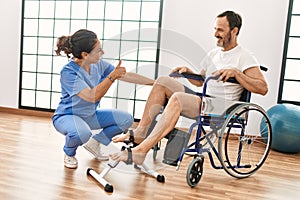 Middle age man and woman having rehab session pedaling sitting on wheelchair at physiotherapy clinic