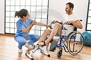 Middle age man and woman having rehab session pedaling sitting on wheelchair at physiotherapy clinic