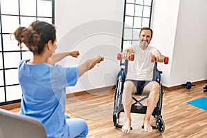 Middle age man and woman having rehab session using dumbbells sitting on wheelchair at physiotherapy clinic