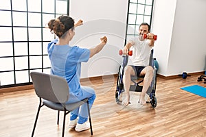 Middle age man and woman having rehab session using dumbbells sitting on wheelchair at physiotherapy clinic
