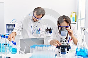 Middle age man and woman partners wearing scientist uniform using laptop and microscope at laboratory