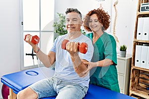 Middle age man and woman wearing physiotherapy uniform having rehab session using dumbbells at physiotherapy clinic