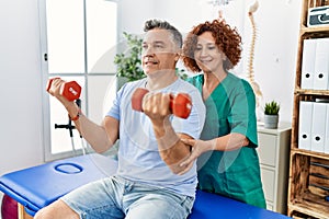 Middle age man and woman wearing physiotherapy uniform having rehab session using dumbbells at physiotherapy clinic
