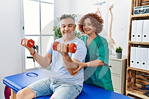 Middle age man and woman wearing physiotherapy uniform having rehab session using dumbbells at physiotherapy clinic