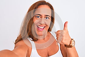 Middle age mature woman taking a selfie photo using smartphone over isolated background happy with big smile doing ok sign, thumb