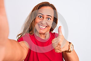 Middle age mature woman taking a selfie photo using smartphone over isolated background happy with big smile doing ok sign, thumb