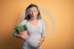 Middle age pregnant woman expecting baby holding plant pot scared in shock with a surprise face, afraid and excited with fear