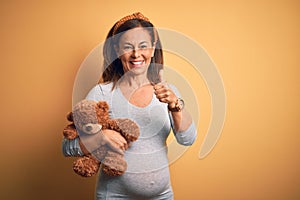Middle age pregnant woman expecting baby holding teddy bear stuffed animal happy with big smile doing ok sign, thumb up with