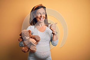 Middle age pregnant woman expecting baby holding teddy bear stuffed animal pointing and showing with thumb up to the side with