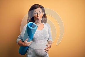 Middle age pregnant woman expecting baby holding yoga mat for healthy lifestyle scared in shock with a surprise face, afraid and