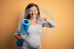 Middle age pregnant woman expecting baby holding yoga mat for healthy lifestyle with surprise face pointing finger to himself
