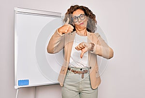 Middle age senior business woman standing on seminar presentation by magnectic blackboard Doing thumbs up and down, disagreement