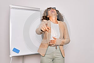 Middle age senior business woman standing on seminar presentation by magnectic blackboard laughing at you, pointing finger to the
