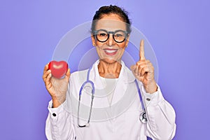 Middle age senior cardiologist doctor woman holding red heart over purple background surprised with an idea or question pointing
