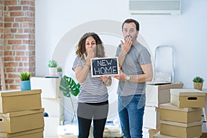 Middle age senior couple holding blackboard moving to a new home cover mouth with hand shocked with shame for mistake, expression