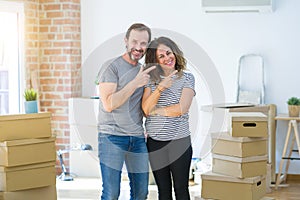 Middle age senior couple moving to a new home with boxes around with a big smile on face, pointing with hand and finger to the