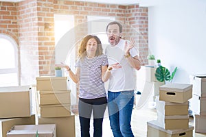 Middle age senior couple moving to a new home with boxes around clueless and confused expression with arms and hands raised