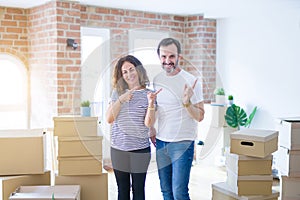 Middle age senior couple moving to a new home with boxes around smiling and looking at the camera pointing with two hands and