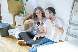 Middle age senior couple moving to a new home using smartphone with surprise face pointing finger to himself