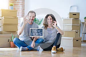 Middle age senior couple sitting on the floor holding blackboard moving to a new home doing ok sign with fingers, excellent symbol