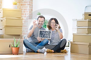 Middle age senior couple sitting on the floor holding blackboard moving to a new home doing ok sign with fingers, excellent symbol