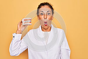 Middle age senior dentist woman holding orthodontics denture with braces aligner scared in shock with a surprise face, afraid and