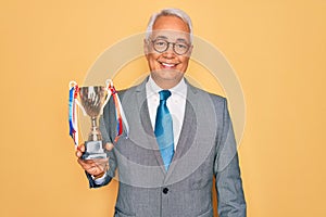 Middle age senior grey-haired business man wearing glasses holding winner trophy with a happy face standing and smiling with a