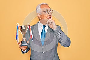 Middle age senior grey-haired business man wearing glasses holding winner trophy serious face thinking about question, very