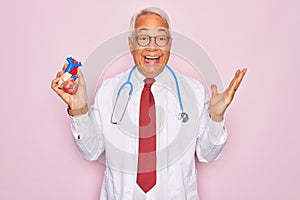 Middle age senior grey-haired cardiologist doctor man holding professional medicine heart very happy and excited, winner