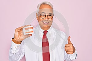 Middle age senior grey-haired dentist man holding prosthesis denture over pink background happy with big smile doing ok sign,